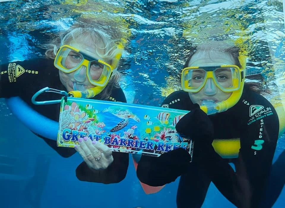 Two snorkelers underwater at the Great Barrier Reef, wearing yellow snorkel masks and holding a colorful 'Great Barrier Reef' sign.