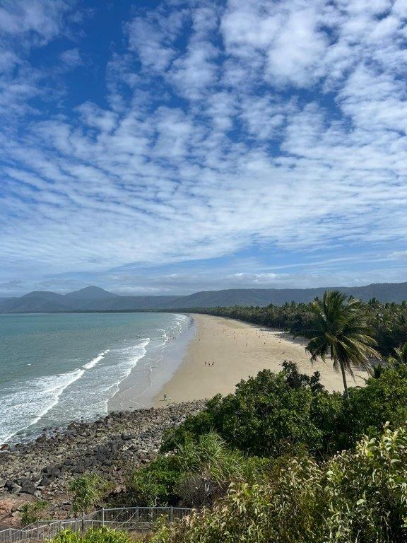 A beach with beautiful clear blue waters and golden sand, and cloudy sky, taken from a vantage point with palm trees and other foliage in view. A mountain silhouette is in the distance.