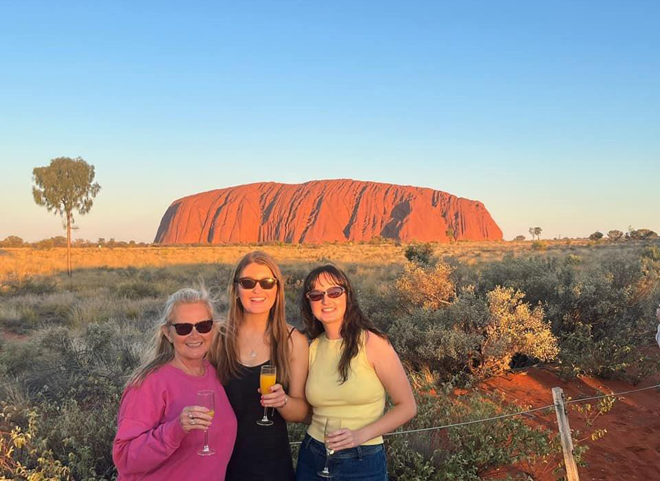 Three people standing in front of Uluru at sunset, smiling and holding drinks, with the iconic red rock formation and a clear blue sky in the background.